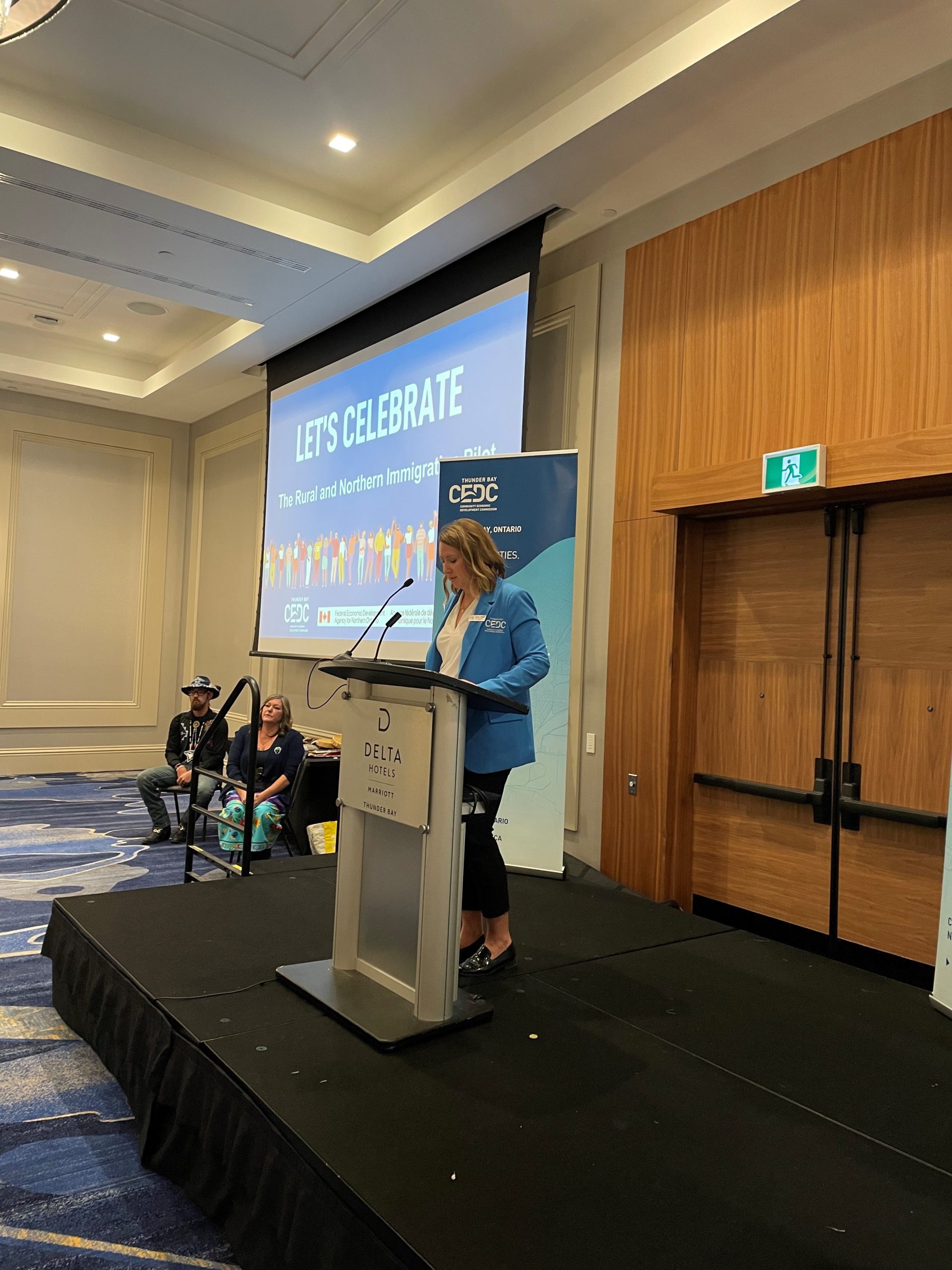 Jamie Taylor -standing on a stage make opening remarks at the Rural and Northern Immigration Pilot event